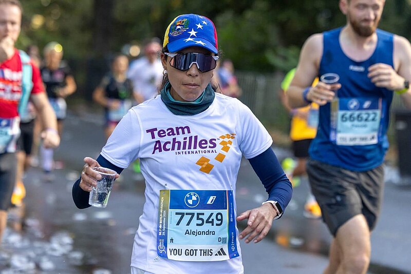 Runner at the BERLIN-MARATHON with a water cup in her hand, headgear, and sunglasses.