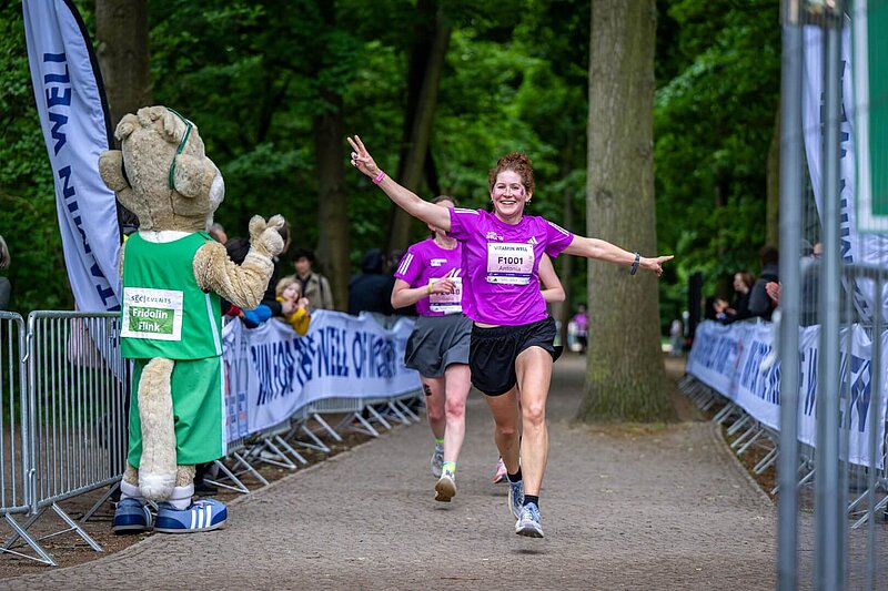 Frau im T-Shirt des 40. VITAMIN WELL Frauenlauf Berlin läuft lachend auf Maskottchen Frido zum abklatschen zu.