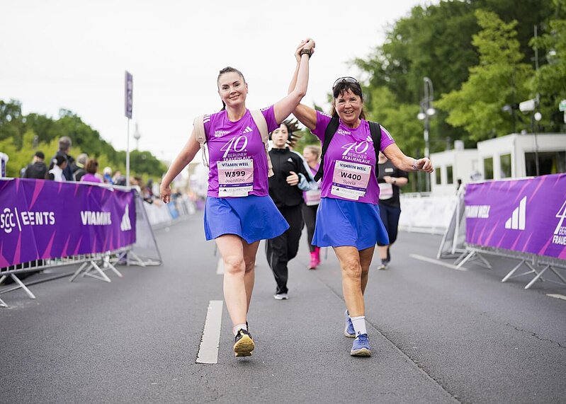 Zwei Frauen in T-Shirts des 40. VITAMIN WELL Frauenlauf Berlin laufen lächelnd Hand in Hand auf der Strecke.