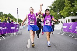 Zwei Frauen in T-Shirts des 40. VITAMIN WELL Frauenlauf Berlin laufen lächelnd Hand in Hand auf der Strecke.