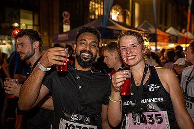 A young woman and a young man toast the photographer with their non-alcoholic beers. They look relaxed and happy and proudly wear their anniversary medals. In the background, there is bustling activity in the finishing area. You can see more participants and Erdinger non-alcoholic tent in the background. © SCC EVENTS | camera 4