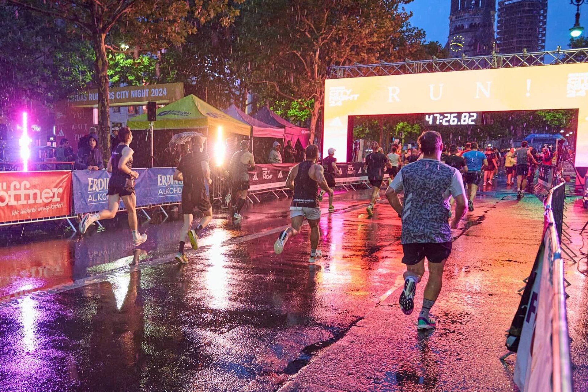 Läufer:innen im Regen in der Dämmerung kurz vor dem leuchtenden Zieltor. Rot-orangene Beleuchtung spiegelt sich auf der Straße und taucht die Szene in warmes Licht.
