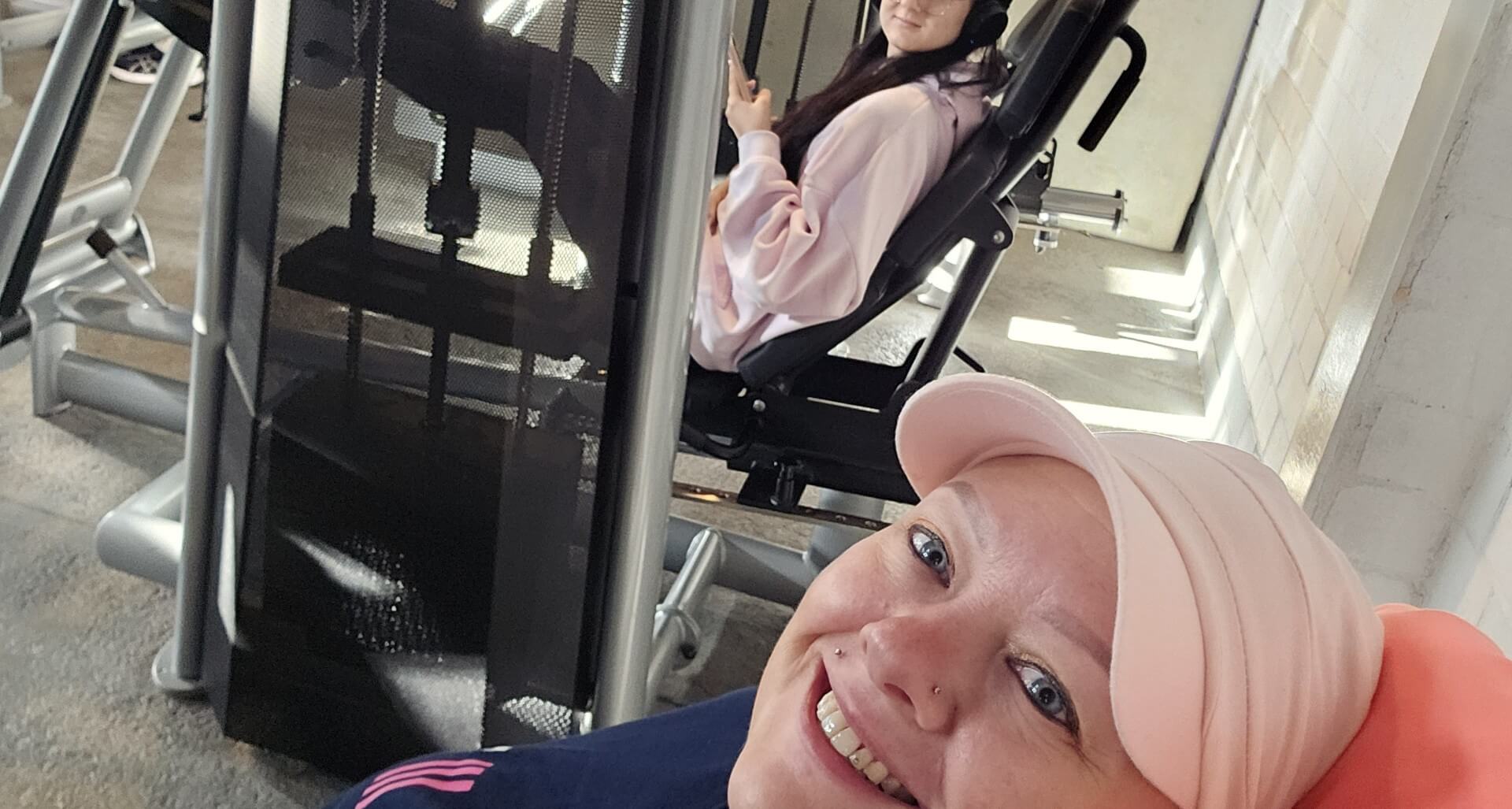 Lächelndes Selfie in einem Raum mit Fitnessgeräten. In einem davon sitzt eine junge Frau.